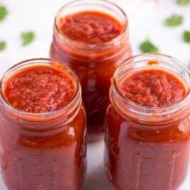 Three mason jars filled with thick, chunky homemade tomato sauce, surrounded by scattered fresh parsley leaves on a white background.