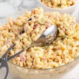 Bowl of macaroni salad with tongs, highlighting creamy dressing coating pasta and crunchy vegetables.