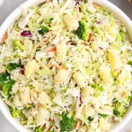 Top-down view of a full bowl of pineapple coleslaw on a marble counter, with pineapple slices and forks nearby.
