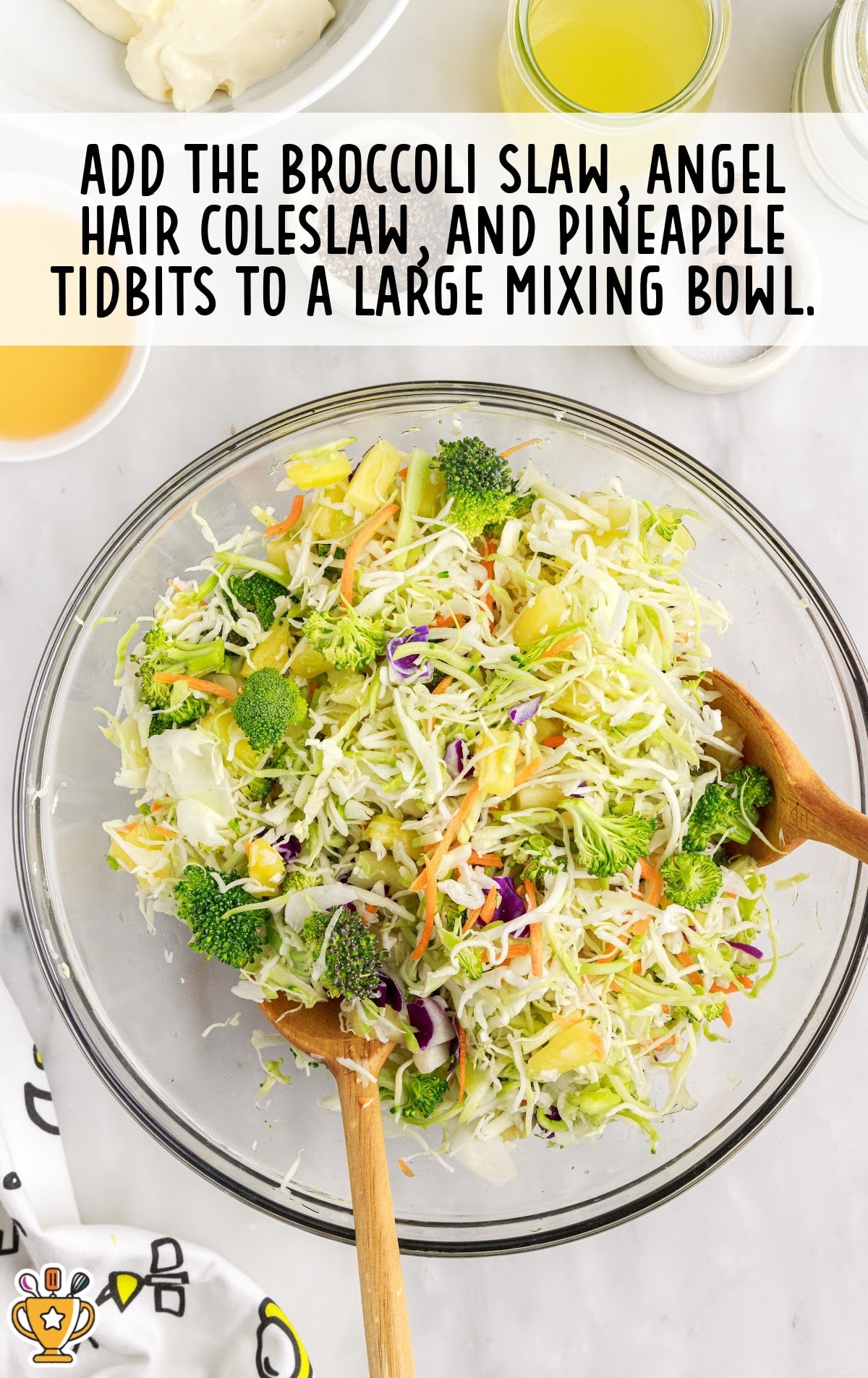 Large glass bowl filled with shredded cabbage, broccoli slaw, and pineapple tidbits being stirred with wooden spoons.