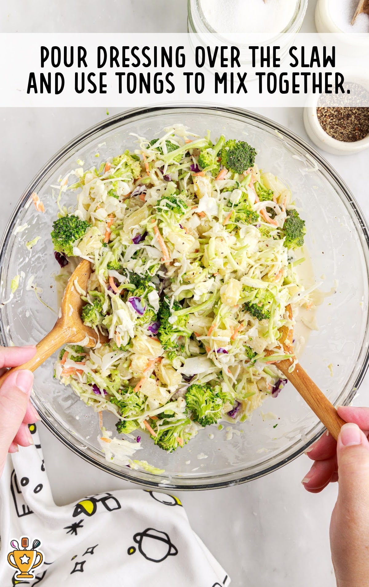 Fresh pineapple coleslaw in a mixing bowl with dressing poured on top, being tossed with wooden tongs.