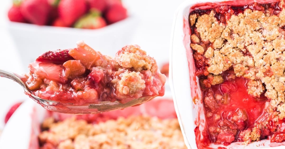 Spoonful of rhubarb strawberry crisp lifted from a white baking dish, with fruit and crumble filling clearly visible.