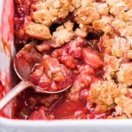 Square image of baked rhubarb strawberry crisp with bubbling fruit filling and a spoon scooping out a portion.