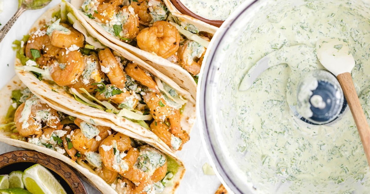 Tacos loaded with shrimp, feta, and cabbage placed beside a large mixing bowl of cilantro lime sauce.
