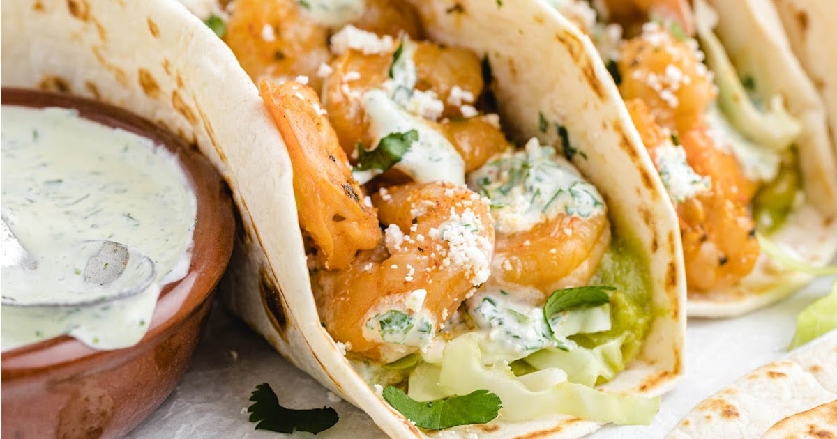 Close-up of shrimp tacos filled with creamy sauce, guacamole, and fresh cilantro next to a bowl of sauce.