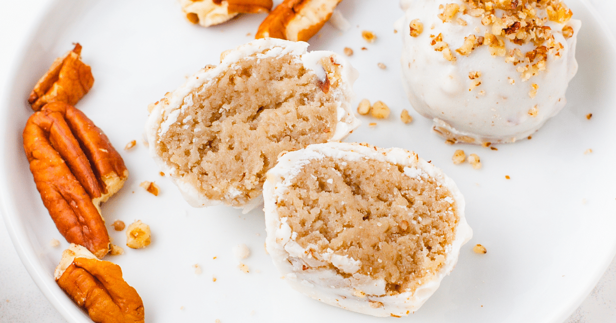 Close-up of butter pecan cheesecake bites on a white plate, with one bite cut in half to show a soft, creamy interior, surrounded by chopped pecans.