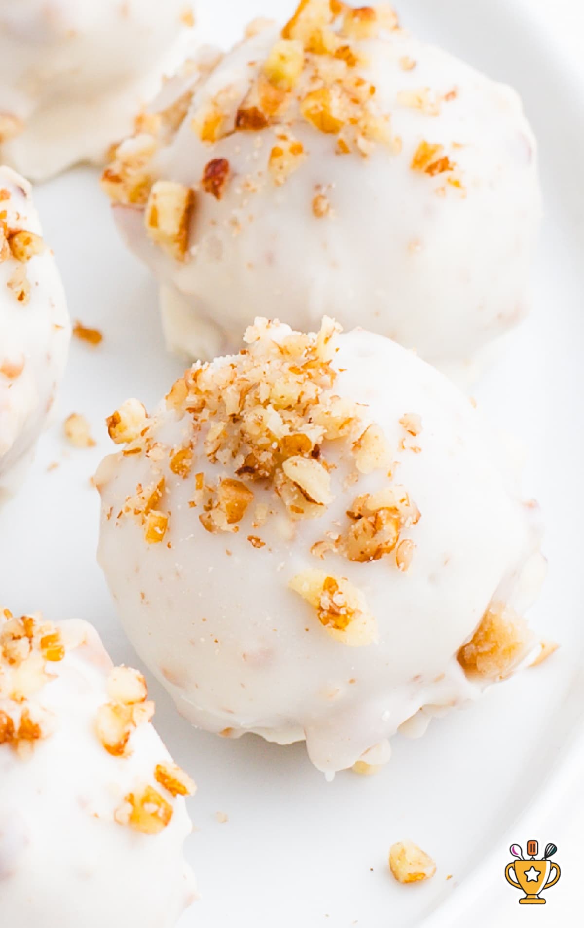 Close-up of butter pecan cheesecake bites topped with chopped pecans and coated in white almond bark, arranged on a white plate.