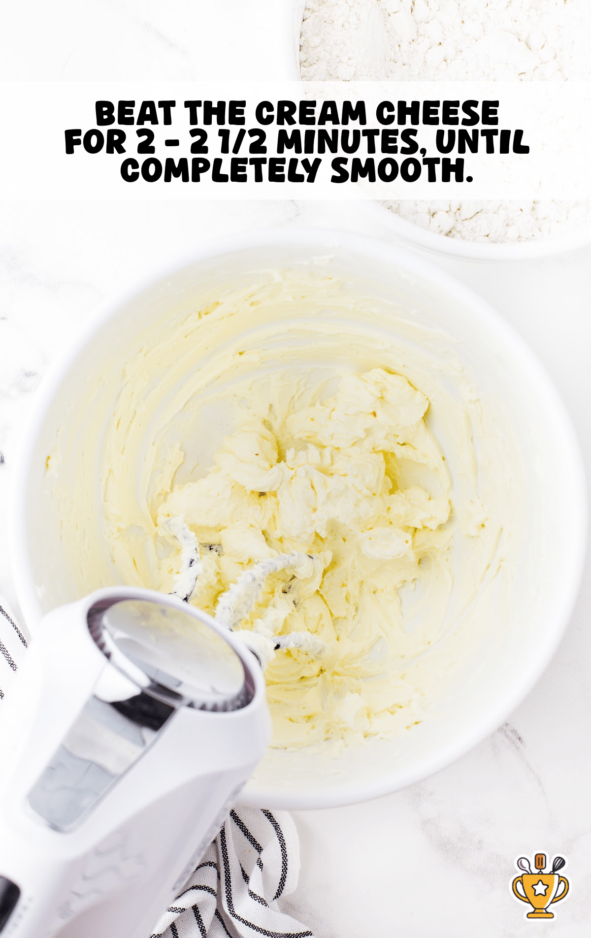 A white mixing bowl with softened cream cheese being whipped with an electric hand mixer until smooth, surrounded by a striped kitchen towel and a bowl of cake mix.