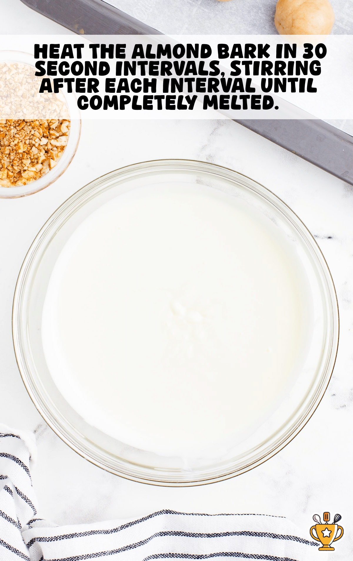 Glass bowl filled with smooth, melted white almond bark next to a small bowl of chopped pecans and a baking tray lined with parchment paper, prepared for dipping cheesecake bites.