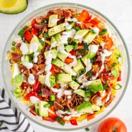 Top-down view of a colorful chicken bacon ranch salad in a glass bowl, featuring chopped avocado, crispy bacon, diced tomatoes, shredded cheese, green onions, and a drizzle of creamy ranch dressing.
