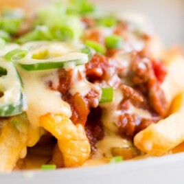Golden fries smothered in beefy chili, creamy cheese sauce, and sliced jalapeños, garnished with fresh green onions on a white plate.