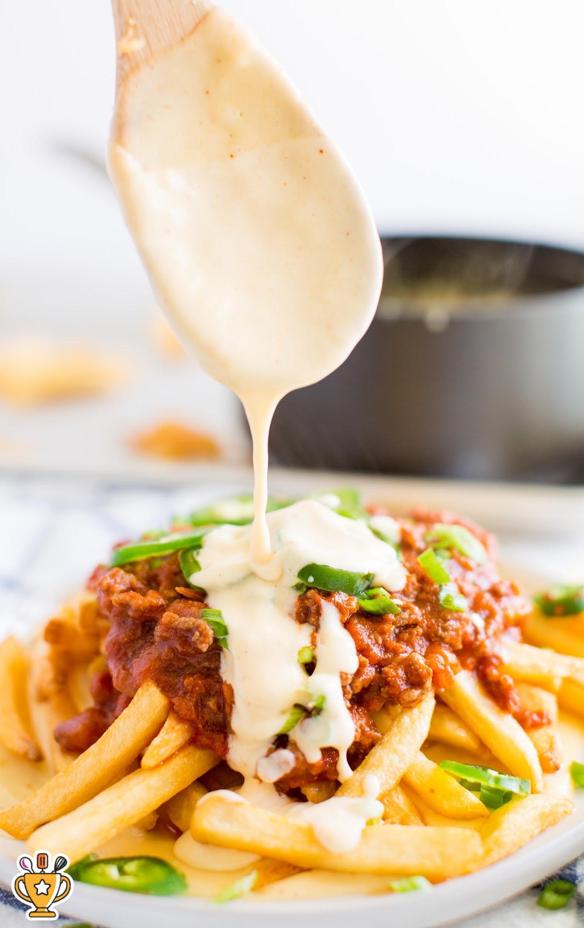 Warm cheese sauce being drizzled from a wooden spoon onto a pile of chili-topped fries with sliced green onions and jalapeños.