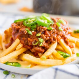 A steaming plate of chili cheese fries with melted cheese, diced onions, and jalapeños on top, served over a soft white napkin.