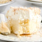 A close-up of a slice of coconut cream pie on a white plate. The pie has a flaky crust, a thick creamy coconut filling, and a generous layer of whipped topping sprinkled with toasted coconut flakes. A bite has been taken out, revealing the rich, textured interior.