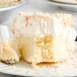A close-up of a slice of coconut cream pie on a white plate. The pie has a flaky crust, a thick creamy coconut filling, and a generous layer of whipped topping sprinkled with toasted coconut flakes. A bite has been taken out, revealing the rich, textured interior.