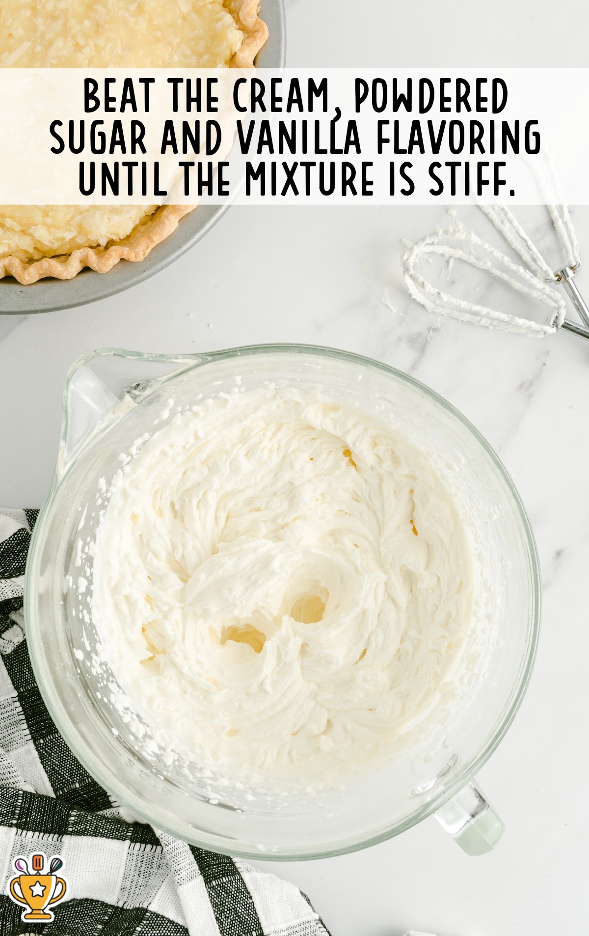 A mixing bowl filled with stiff whipped cream next to a partially filled pie crust and beaters on a white countertop