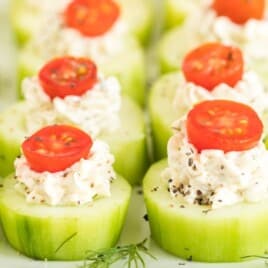Crisp cucumber rounds filled with creamy herb spread and topped with cherry tomato slices, garnished with cracked black pepper.