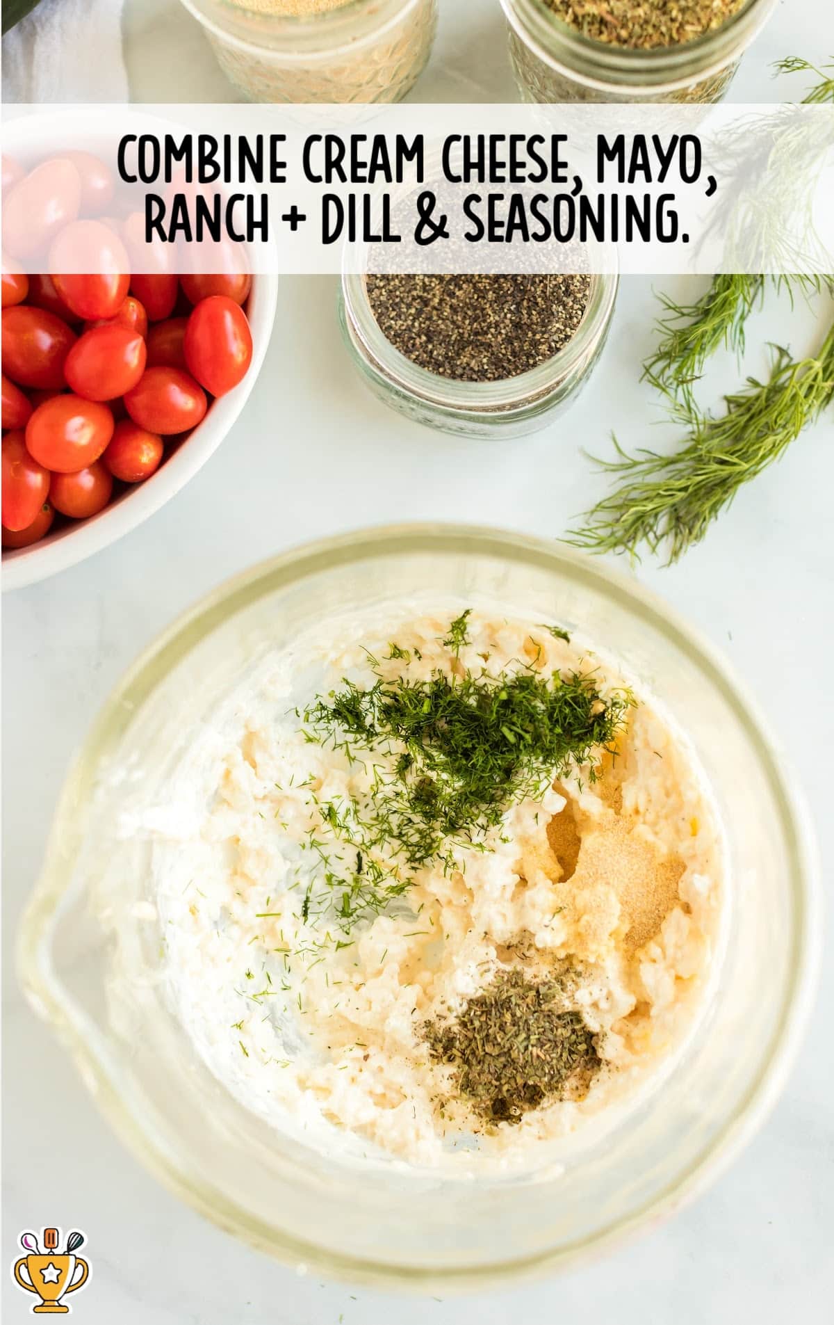 Mixing bowl with cream cheese, mayo, dill, ranch mix, and seasonings surrounded by cherry tomatoes and jars of spices.