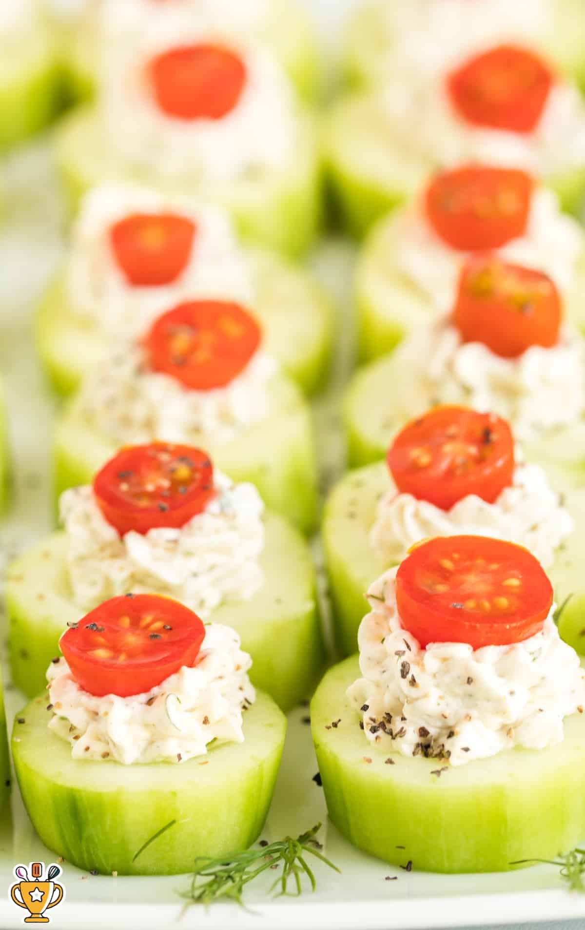 Close-up of cucumber bites garnished with black pepper and dill, topped with cherry tomato slices and neatly aligned.