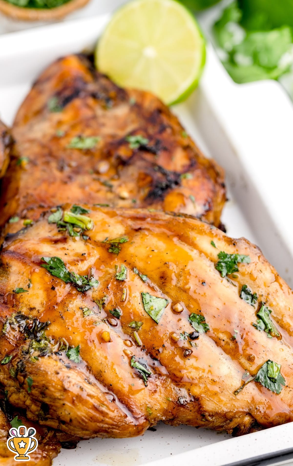 Juicy grilled chicken breasts layered on a white tray with fresh cilantro and lime slices on the side.