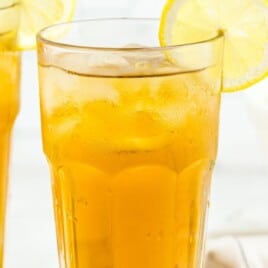 Two glasses of iced tea with lemon slices, one filled with ice, sitting on a white surface with lemons in the background.