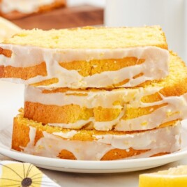Three slices of lemon pound cake stacked neatly on a white plate, with icing glaze dripping down the sides, set on a white table with fresh lemon halves and a floral kitchen towel.