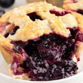 Close-up of a mini blueberry pie on a white dish, bursting with thick, glossy blueberry filling and topped with a sugar-dusted crust.