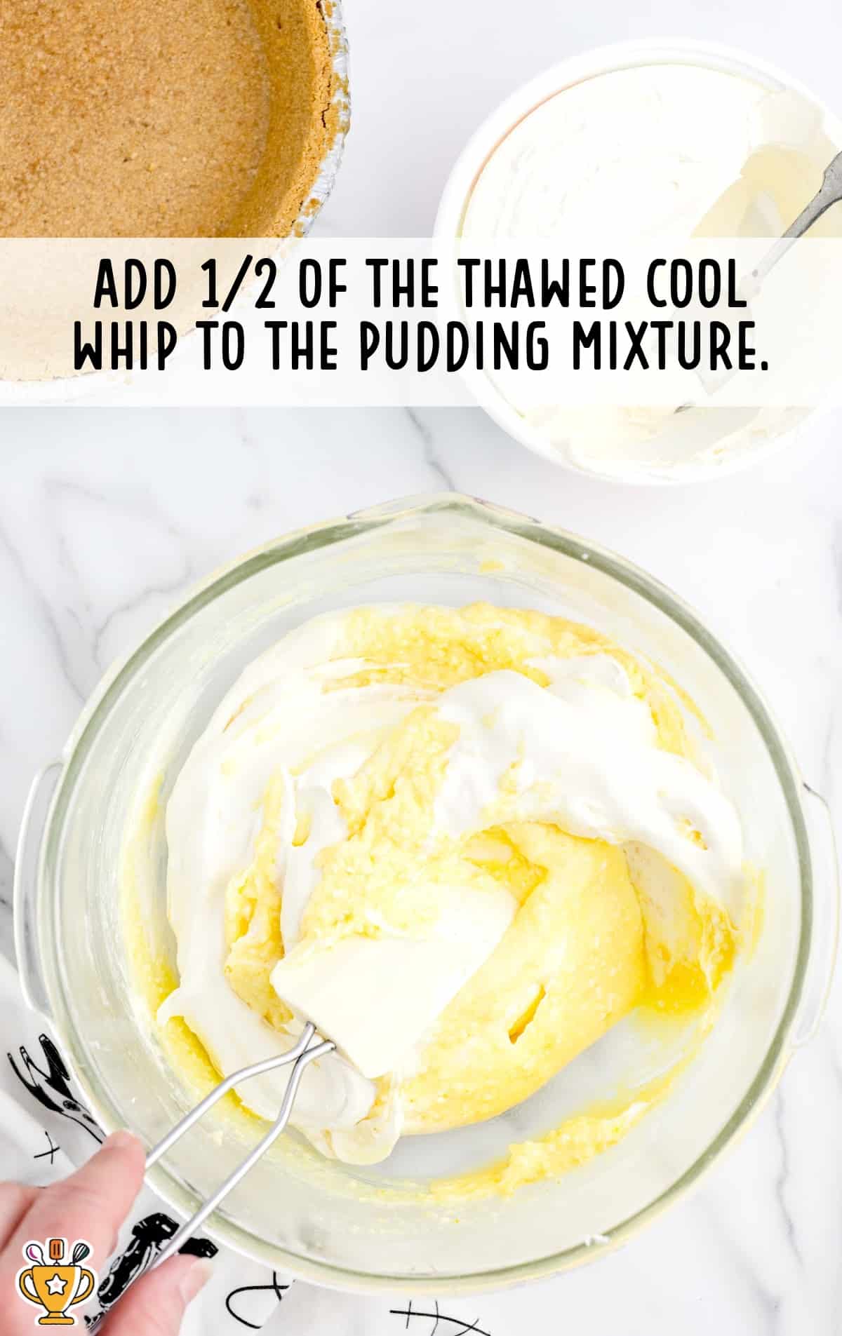 Cool Whip being folded into the yellow pudding mixture using a spatula, with the graham crust and Cool Whip container in view