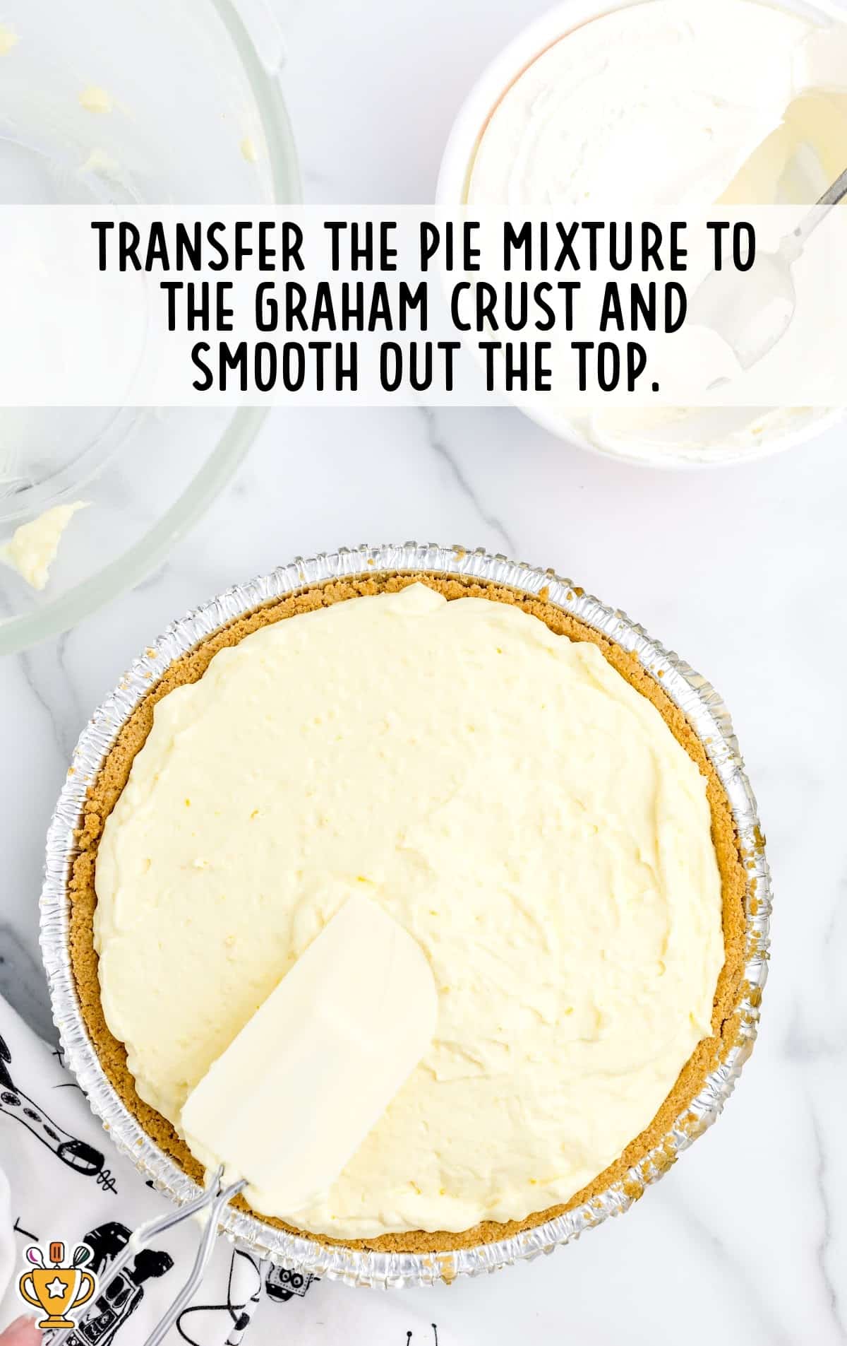 The creamy pie filling being spread into a graham cracker crust using a white spatula, ready for the next step