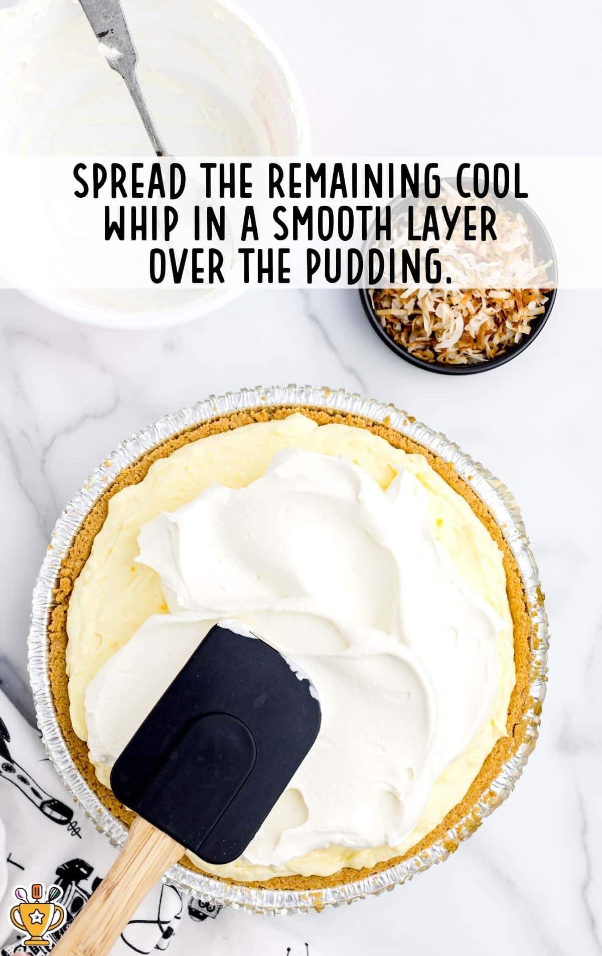 A black spatula smoothing a thick layer of Cool Whip over the top of the pudding layer in the pie crust.
