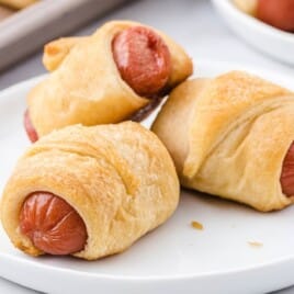 A full view of a plate of pigs in a blanket, showing several golden-brown, puffed-up dough-wrapped hot dogs ready to be served.