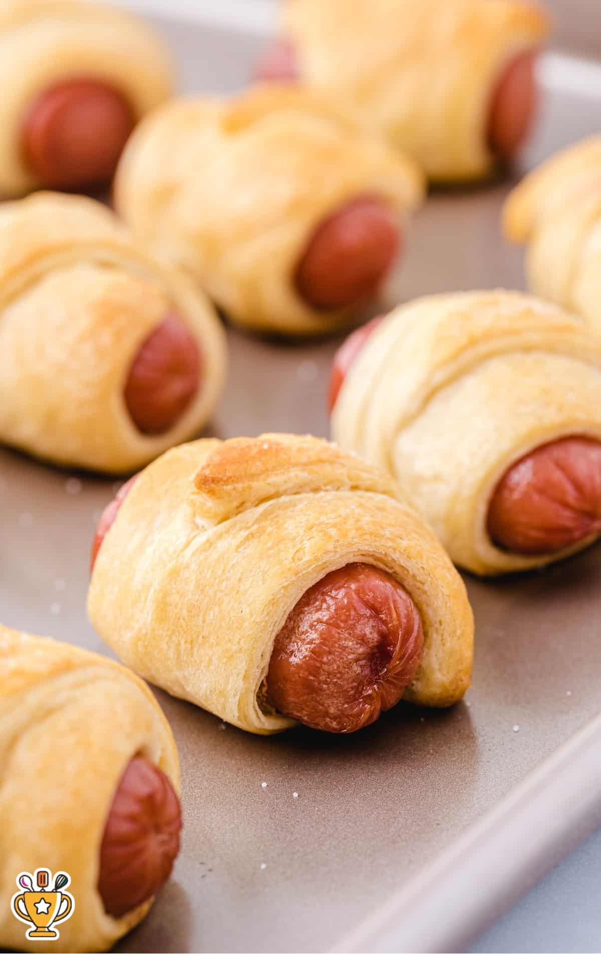 A close-up of freshly baked pigs in a blanket on a baking tray, showcasing the golden, crispy exterior of the dough wrapped around hot dogs.