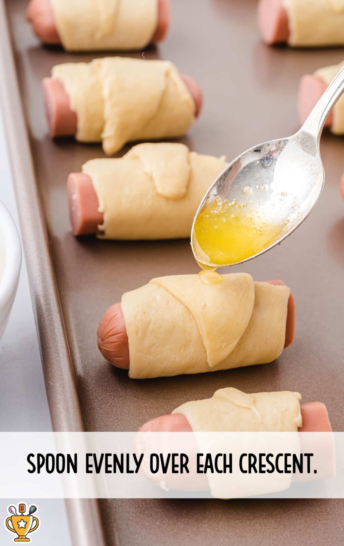 A spoon is drizzling melted butter evenly over crescent-wrapped cocktail wieners on a baking sheet.