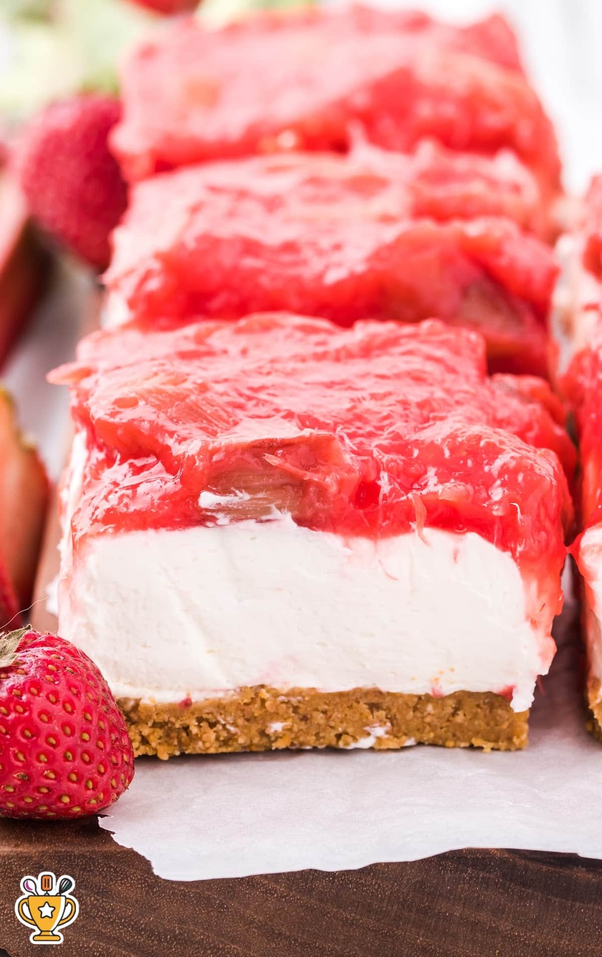 A close-up of a slice of strawberry rhubarb cheesecake topped with a generous amount of homemade rhubarb and strawberry compote. The cheesecake is displayed on a white plate with fresh strawberries in the background.