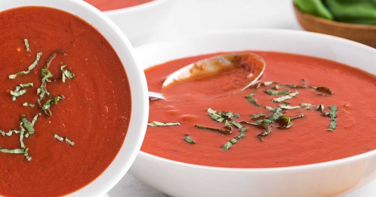 A close-up of tomato soup in a white bowl with a spoon resting inside, garnished with freshly cut basil leaves.