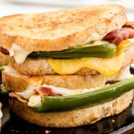 Close-up of a toasted grilled cheese sandwich stacked with jalapeño halves, bacon, and melted cheese on a hot skillet.