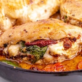 A close-up of stuffed chicken breasts in a skillet, with sun-dried tomatoes, spinach, and melted cheese, simmering in sauce.