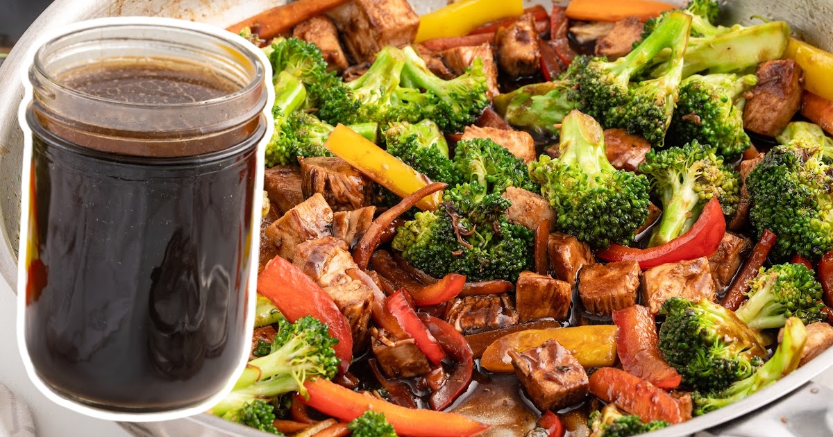 A jar of dark brown stir fry sauce in front of a colorful vegetable stir fry with broccoli, red and yellow bell peppers, tofu cubes, and carrots in a skillet, all coated in glossy sauce.