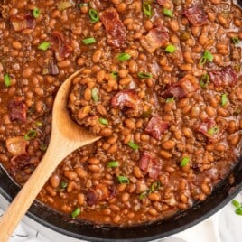 Close-up of baked beans with ground beef in a skillet, topped with crispy bacon and sliced green onions, with a wooden spoon scooping a hearty portion.
