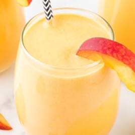 Glass of peach banana smoothie garnished with a fresh peach slice and black striped straw, surrounded by more smoothies and peach slices.