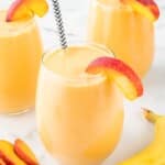 Peach banana smoothie in a glass with peach garnish and striped straw, placed in front of additional smoothies. Smooth texture visible through glass.