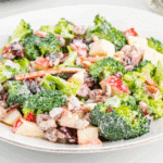 A fresh and colorful broccoli apple salad served on a white plate. The salad features bright green broccoli florets, crisp red apple chunks, shredded carrots, dried cranberries, and chopped pecans, all coated in a creamy dressing.