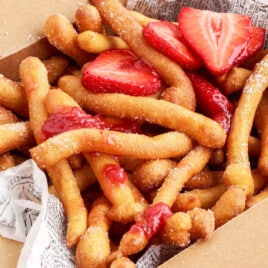 Box of golden funnel cake fries coated with sugar, garnished with fresh sliced strawberries and a generous drizzle of strawberry sauce.
