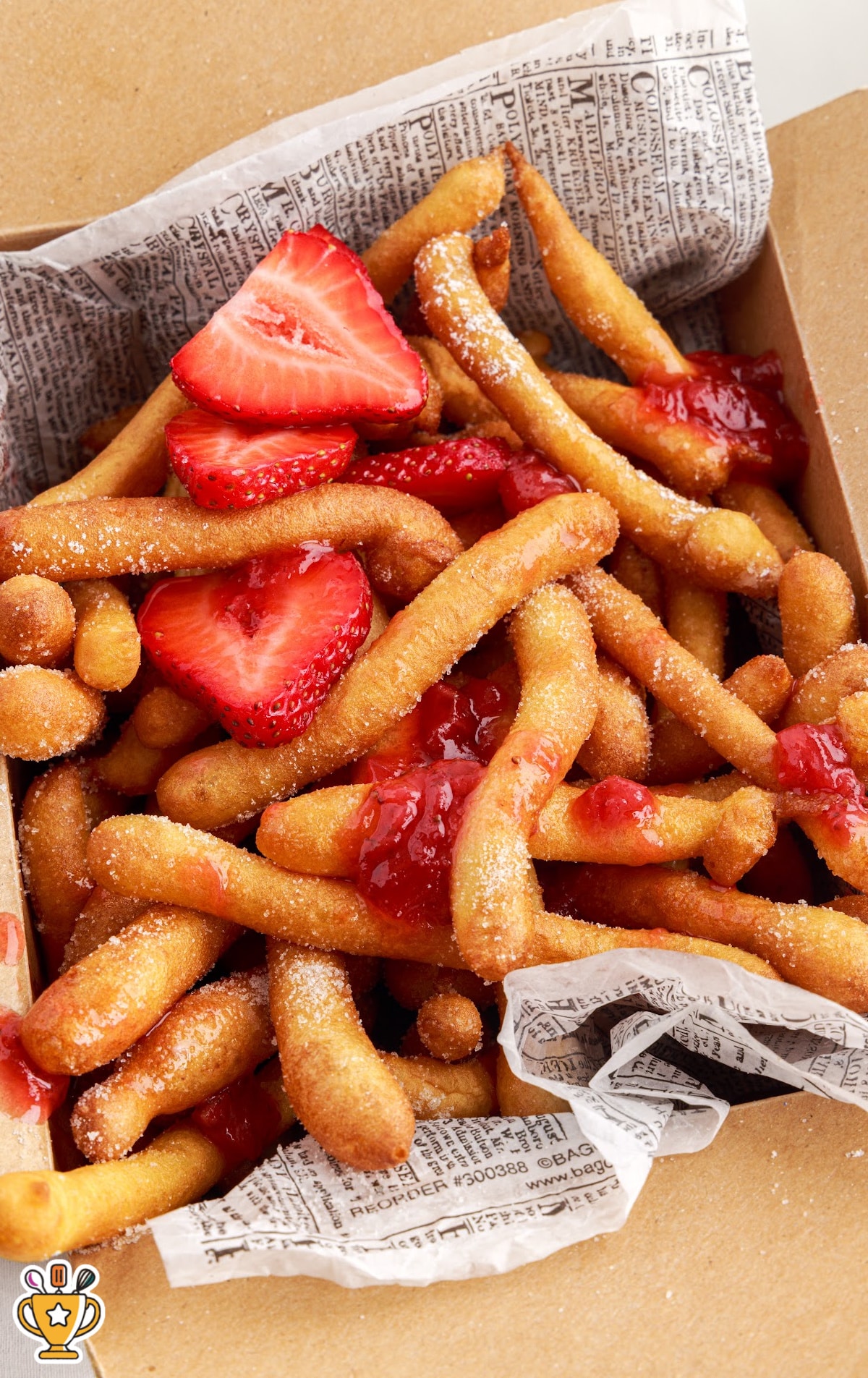 Close-up of golden funnel cake fries in a cardboard tray lined with parchment and topped with sliced strawberries and strawberry sauce.