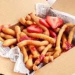 Box of funnel cake fries sprinkled with sugar, topped with strawberries and strawberry sauce, ready to serve in a paper-lined tray.