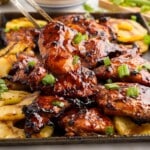 Tongs lifting a piece of glossy, grilled Huli Huli chicken from a tray surrounded by grilled pineapple and garnished with green onions.