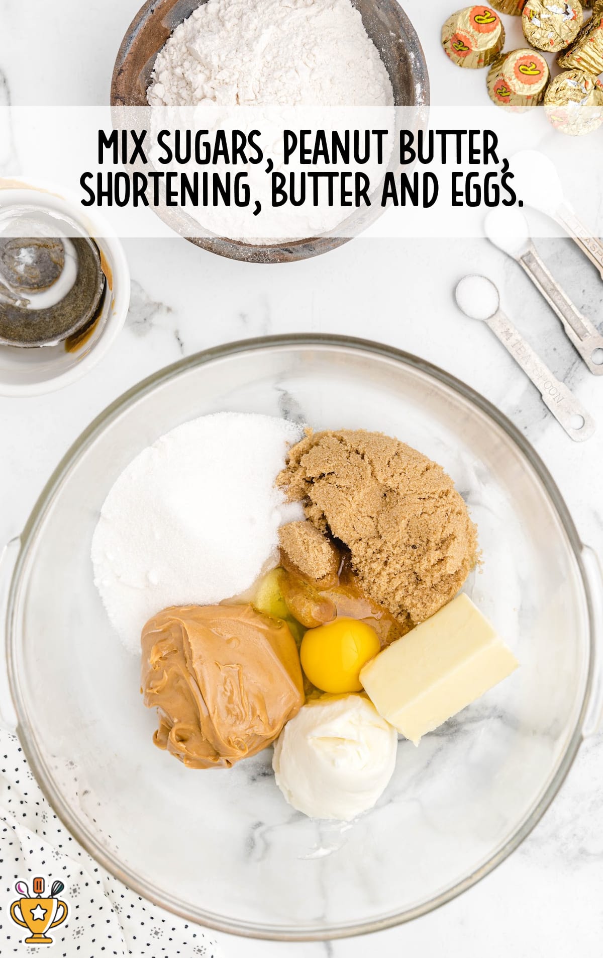 Glass mixing bowl with peanut butter, brown sugar, white sugar, egg, butter, and shortening on a marble counter with ingredients nearby.