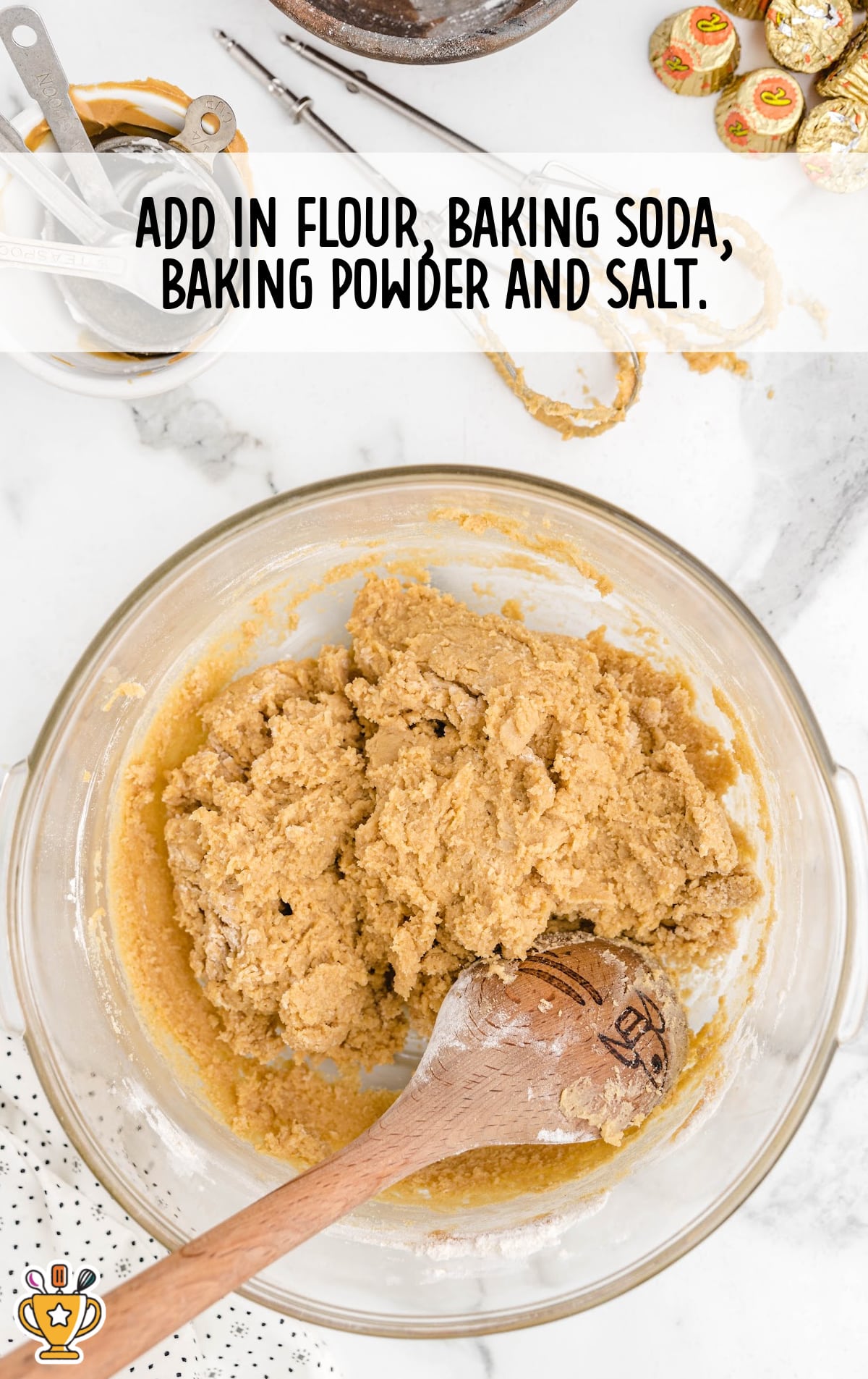 Creamy peanut butter cookie dough in a glass bowl with wooden spoon, surrounded by baking tools and ingredients on a white marble surface.