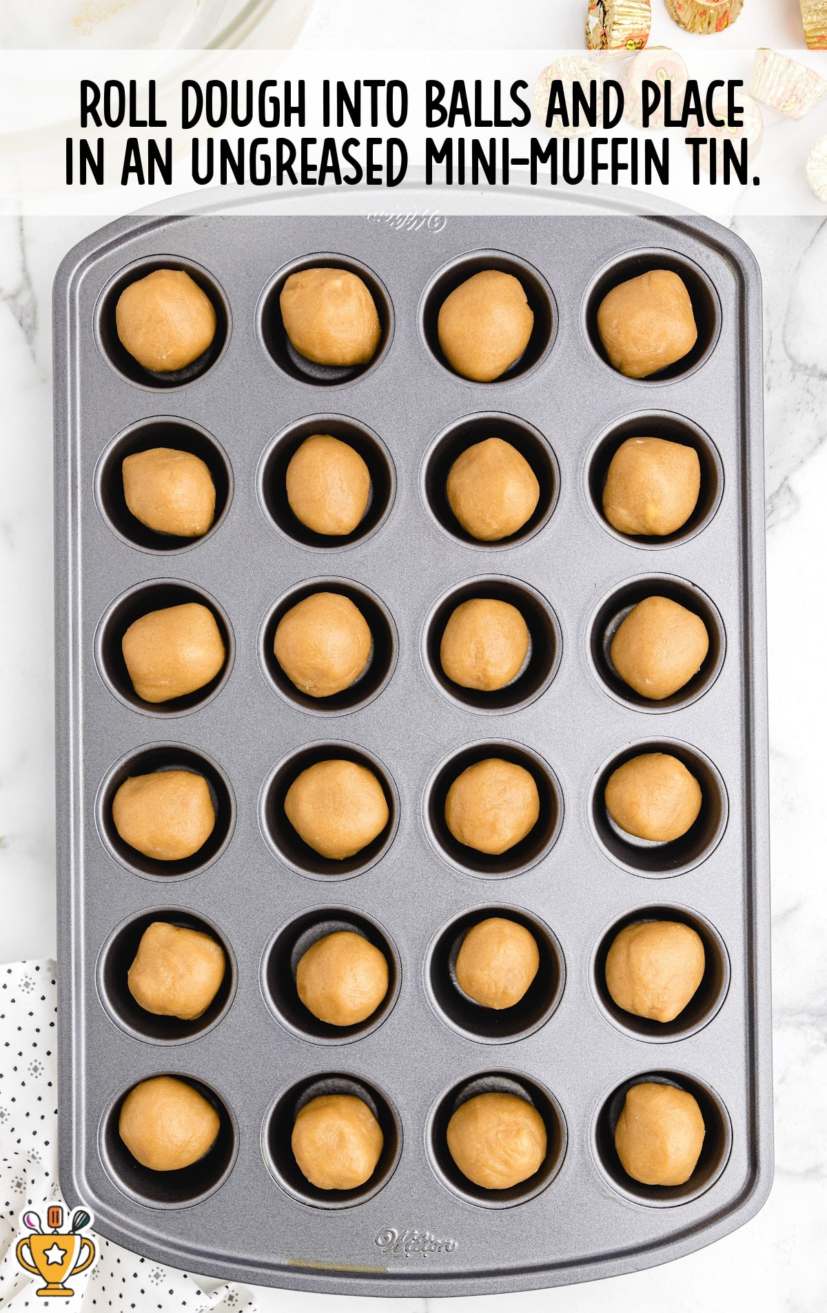 Mini muffin tin filled with evenly rolled peanut butter dough balls, ready to bake on a white marble countertop.