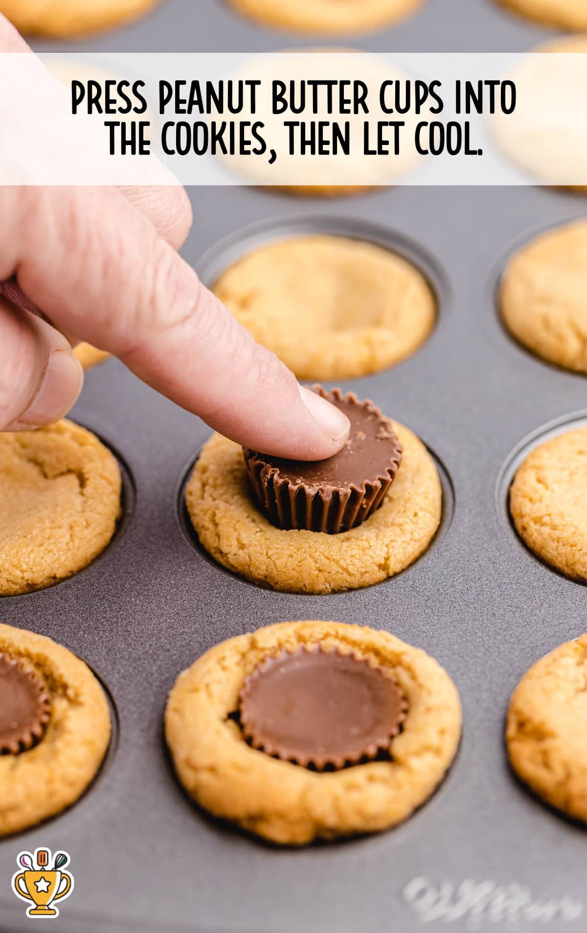 A hand pressing a peanut butter cup into a freshly baked cookie inside a mini muffin tin, cookies still in the pan.